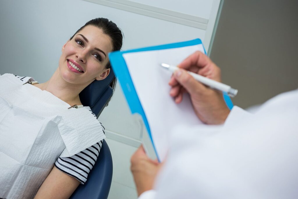 Befund wird dokumentiert Zahnarzt notiert den Befund auf einem Klemmbrett, die Patientin sitzt entspannt im Behandlungsstuhl