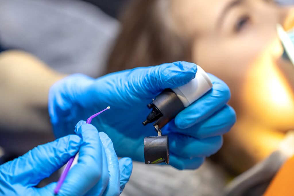 Mikrobürstchen für die Zahnhalsbehandlung Behandschuhte Hände halten ein Mikrobürstchen und einen Applikator mit Dentalmaterial, im Hintergrund unscharf eine Patientin