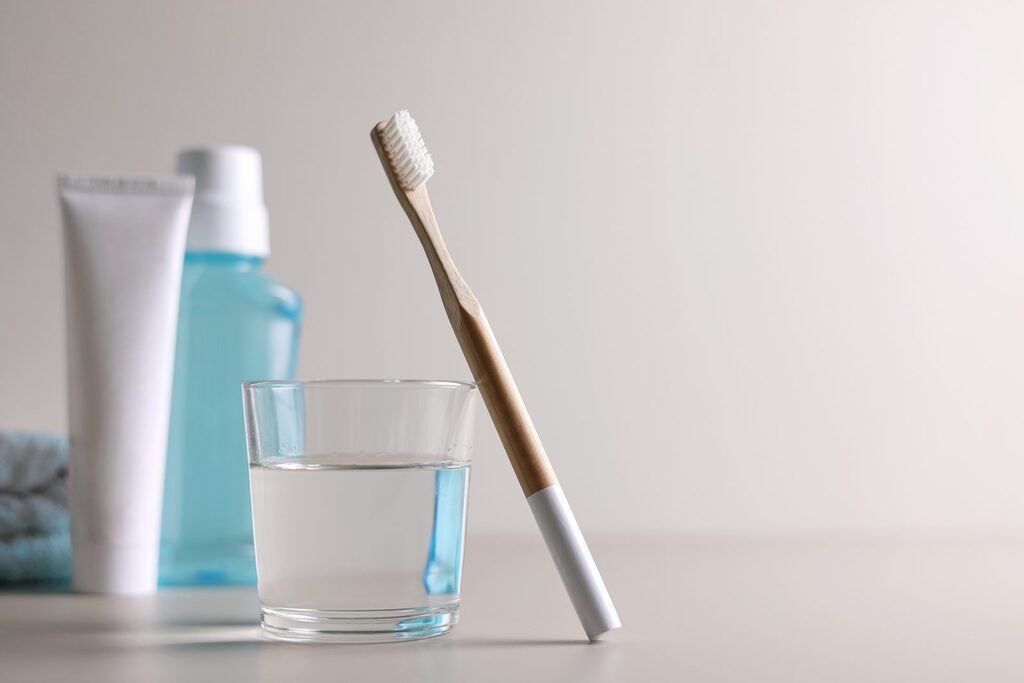Ausspülen nach dem Erbrechen Glas Wasser, blaue Mundspülung und Zahnpastatube neben einer Bambus-Zahnbürste mit hellen Borsten auf weißem Tisch