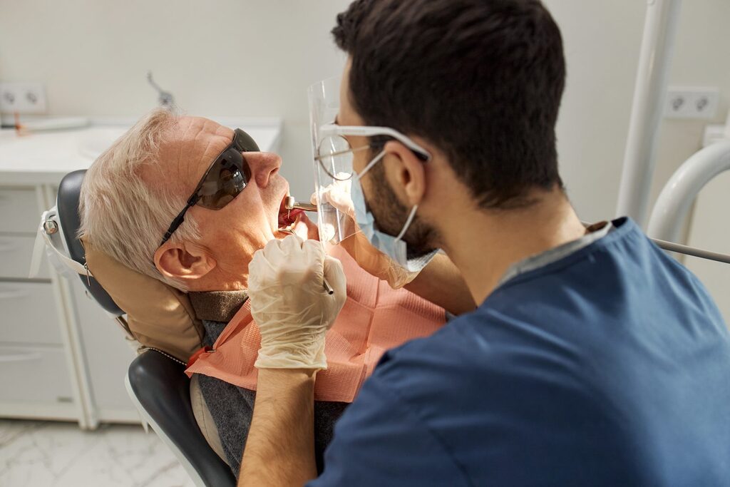 Kontrolle in der Implantatnachsorge Zahnarzt mit Mundschutz untersucht die Zähne eines älteren Patienten mit Schutzbrille mithilfe zahnärztlicher Instrumente