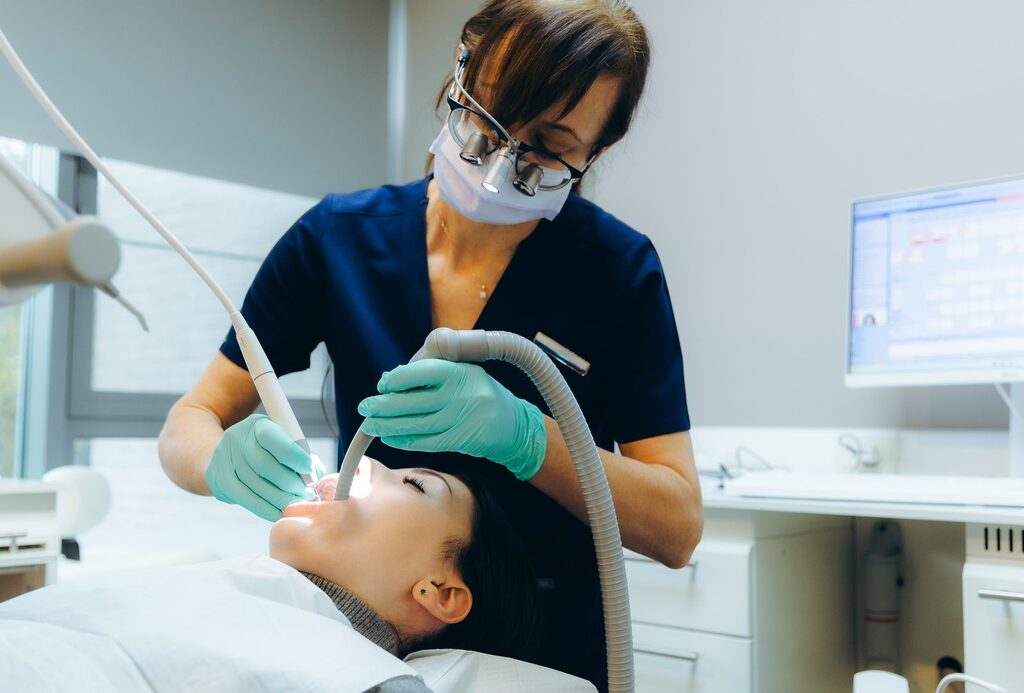 Zahnsteinentfernung in der Praxis Zahnmedizinische Fachkraft mit Lupenbrille reinigt die Zähne einer Patientin mit Handinstrument und Absaugschlauch