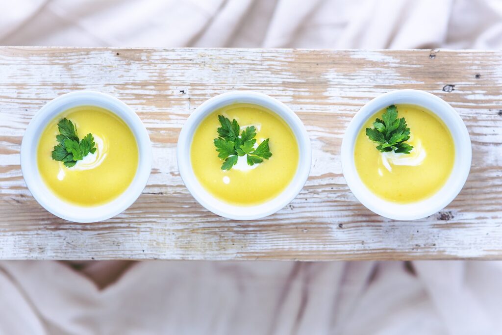 Cremesuppe als weiche Kost Drei weiße Schälchen mit cremiger, gelber Gemüsesuppe und Petersilie auf hellem Holz als weiche Kost nach der Zahnentfernung