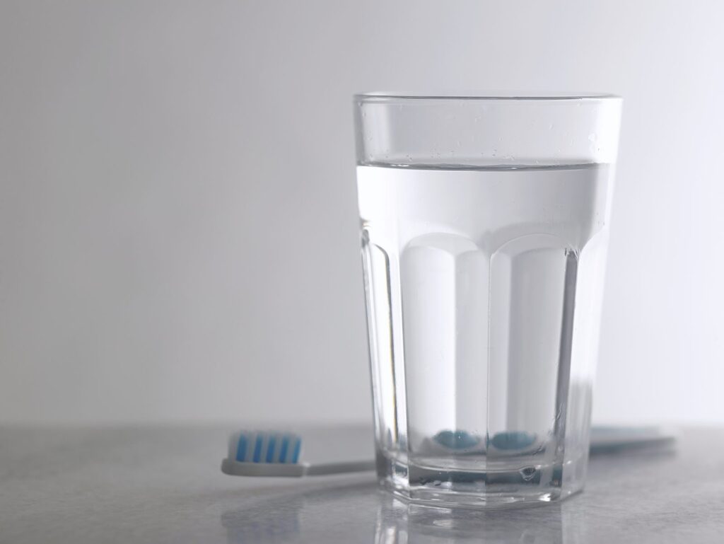 Zahnpflege rund um die Wunde Weisheitszahn OP Nachsorge: Zahnbürste mit blau-weißen Borsten liegt neben einem Glas Wasser auf grauer Steinplatte